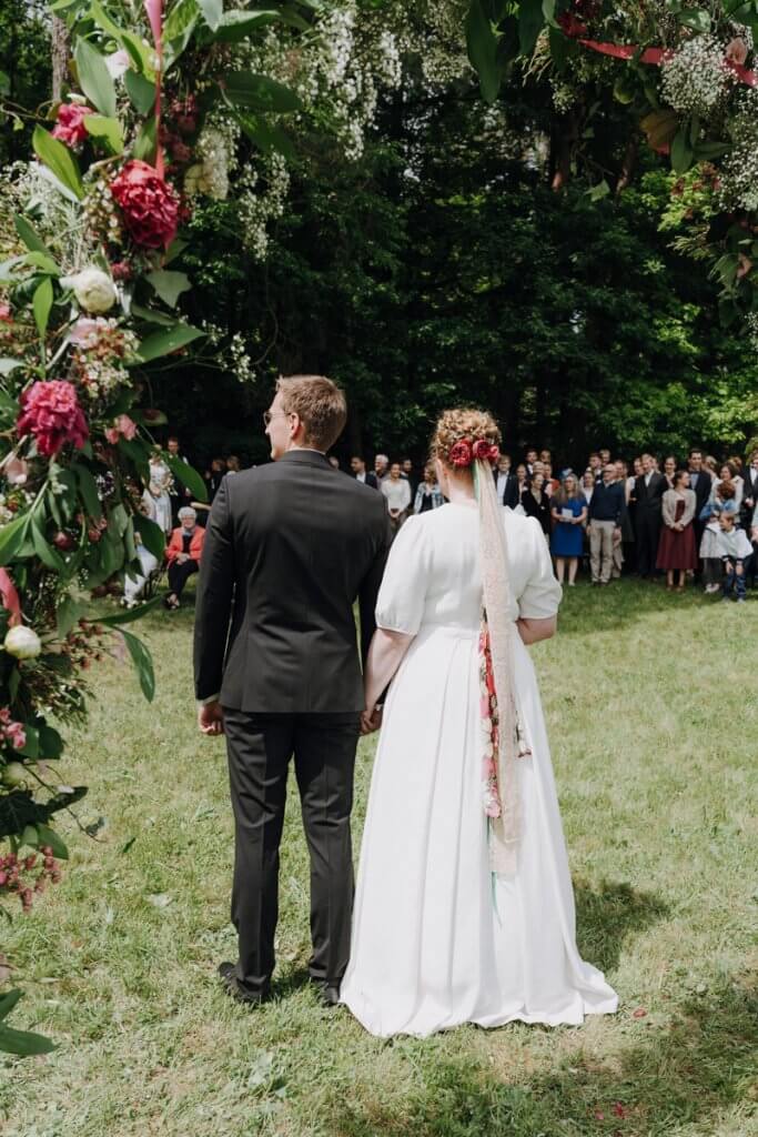 Ein Brautpaar steht unter dem Traubogen vor ihre Gästen bei ihrer Hochzeit